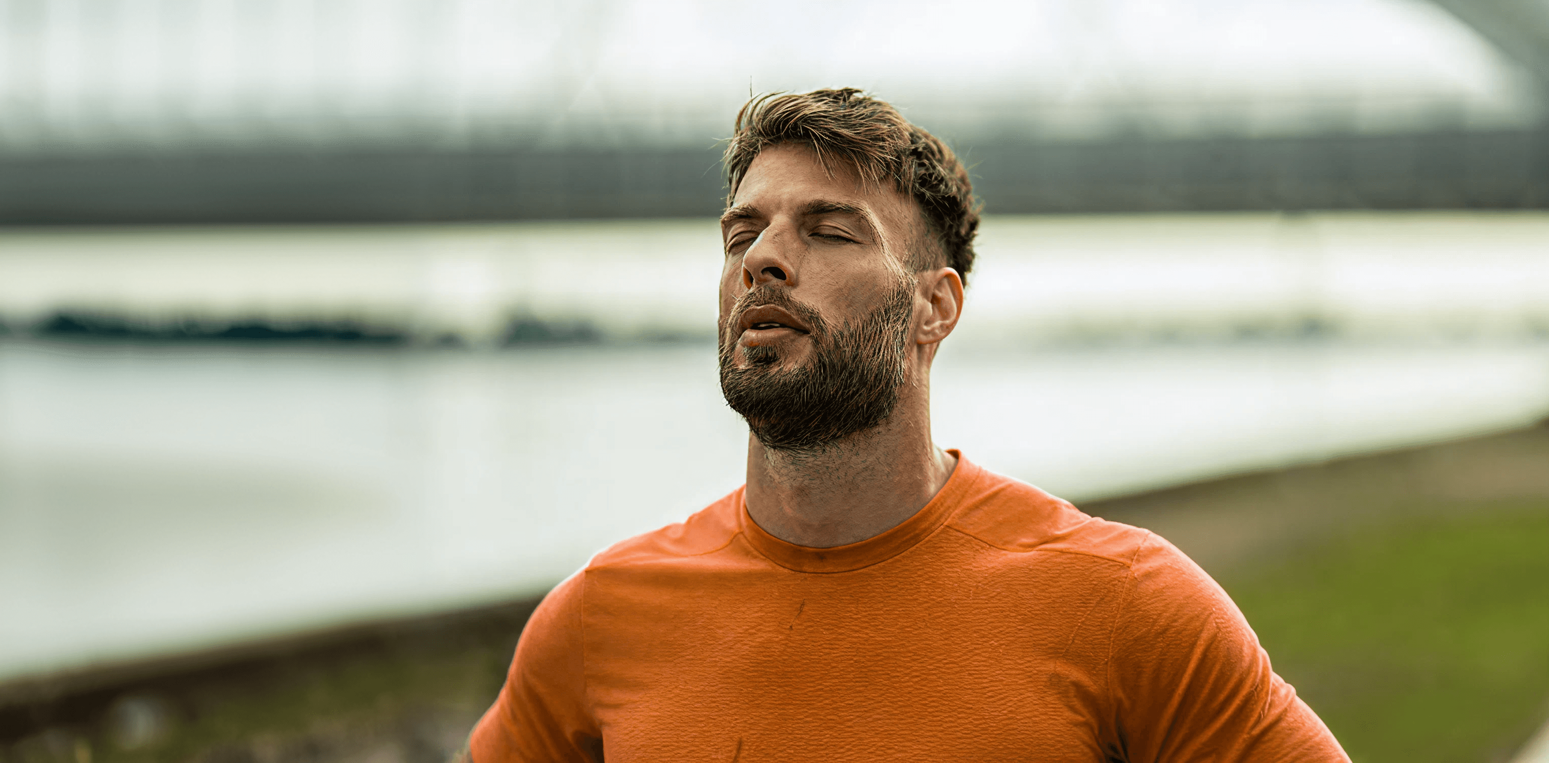 full-bleed photo of a bearded man in an orange athletic shirt, eyes closed, outdoors by a blurred waterfront